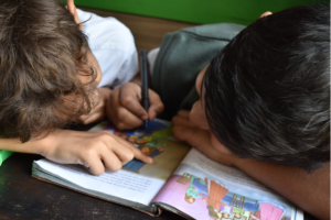 Zwei Kinder Arbeiten lehnen dicht über einer Zeitschrift, eines der Kinder schreibt oder malt mit einem Stift, das andere zeigt auf eine Textstelle, lesen lernen