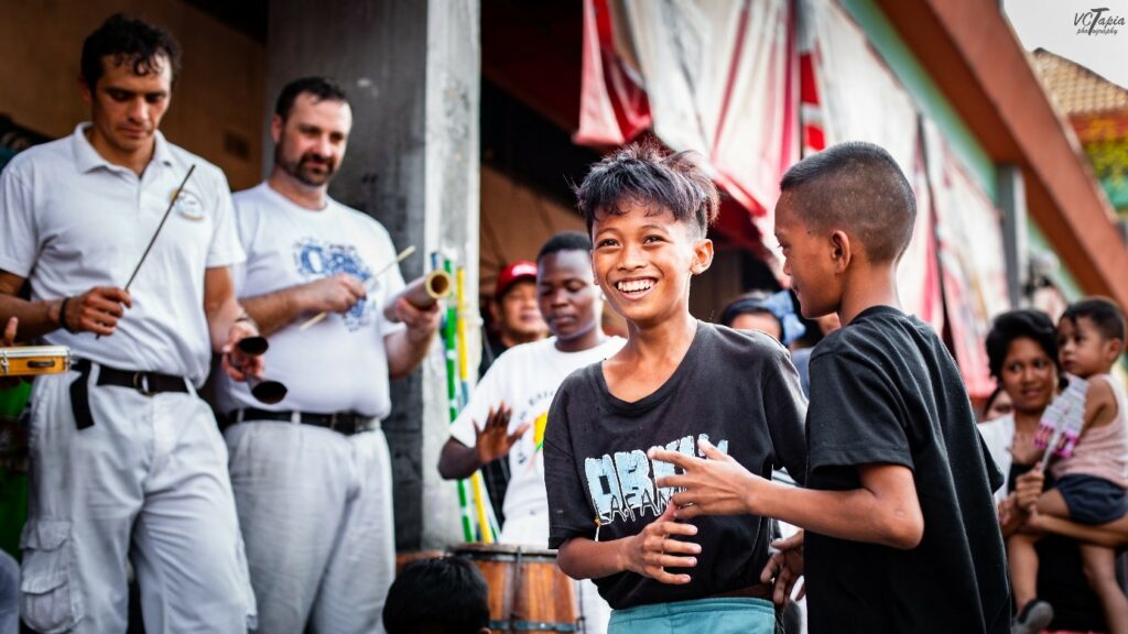 Der Sport entsteht aus den Tänzen und der Musik der Sklav*innen, die von der Westküste Afrikas nach Brasilien verschleppt werden. Die Geschichte der Capoeira ist schlecht dokumentiert, da man sie vor allem mündlich weitergab. Sie hängt jedoch mit dem Widerstand gegen die Gewalt der Kolonialist*innen zusammen. Anfangs praktizieren die Sklav*innen Capoeira zur Selbstverteidigung, maskiert als Tanz, denn die Kolonialist*innen verbieten ihnen das Ausüben von Kampfsportarten.