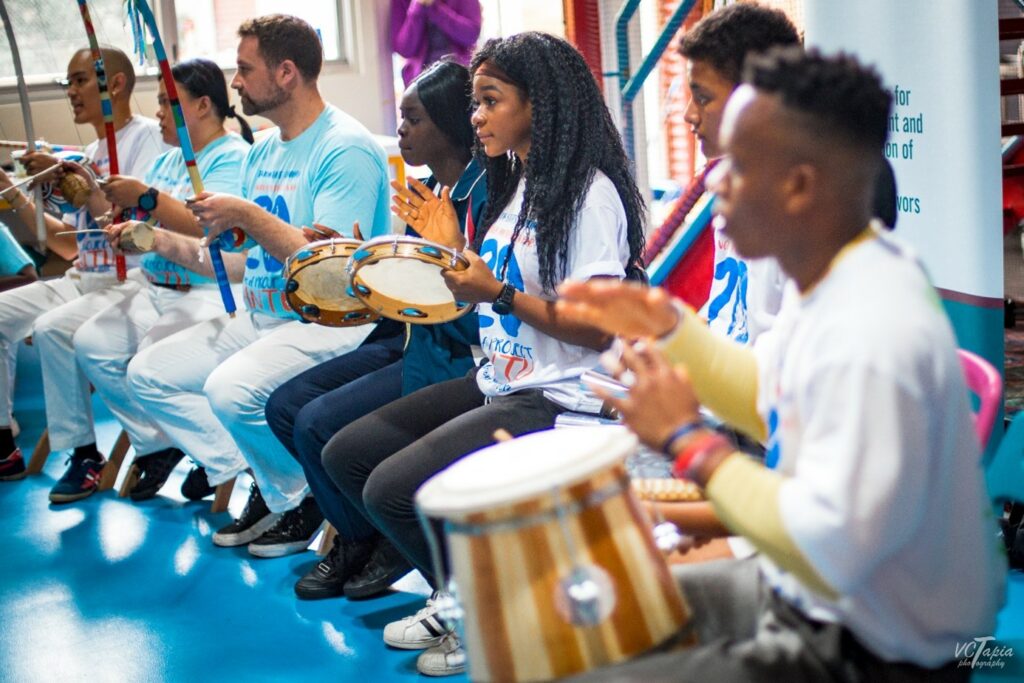 Die Bateria, also das Musikensemble der Capoeira, wird angeführt von der Berimbau. Ihr seht sie weiter hinten im Bild: ein langer Holzbogen, auf den eine Saite aufgespannt wird. Unten befindet sich der Resonanzkörper, die Kalebasse. Der*die Spieler*in der Berimbau darf als Einzige*r den Rhythmus der Musik verändern. Unterstützt wird die Berimbau von der Pandeiro, einem Tamburin, und der Atabaque, einer hohen Trommel (hier vorne im Bild). Abgesehen von diesen Hauptinstrumenten enthalten manche Baterias auch eine Agogo, ein glockenähnliches Instrument, oder ein Rego-Rego. In den Liedern der Capoeira steckt viel Geschichte: Sie erzählen vom Elend der Sklaverei, von der Befreiung der Schwarzen Bevölkerung oder beziehen sich auf Spirituelles wie den Candomblé, eine brasilianische Religion mit Ursprung in Westafrika.